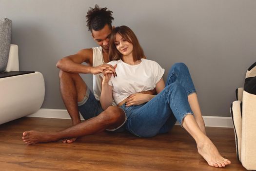 A multiracial couple, a guy and a girl in white t-shirts and blue jeans, sit on the floor at home against a gray wall, hugging, holding hands. Copy space