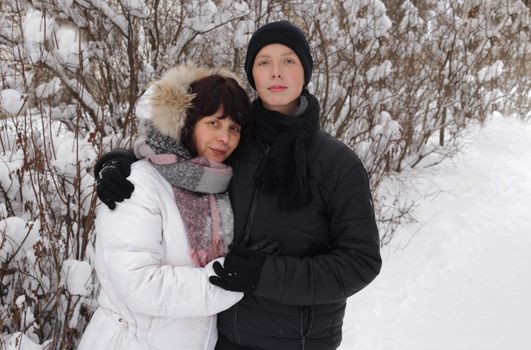 A woman in white winter clothes and her son in a black jacket and hat and glove in front of a snow-covered bush. family outdoors. cheerful mommy with her child.