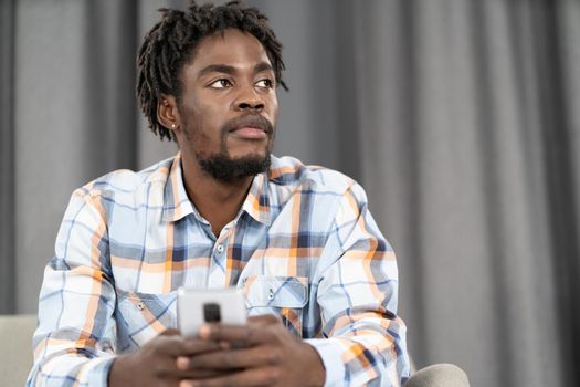 Thoughtful African American man use smartphone checking social media or updating information. Portrait of young man holding smartphone sitting on the sofa. Social media concept.