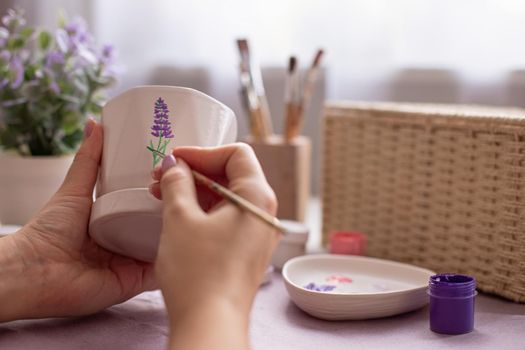 Female hands paints with a brush a sprig of a lavender flower, on a white ceramic pot, with a brush. There are brushes, jars of paints and a palette on the table. Close-up.