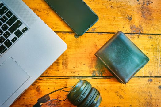 Top view of laptop, headphones, smartphone, and leather wallet on wooden table. Freelancer's work surface. Modern devices for business jobs and entertainment.