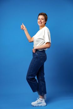 Young happy cheerful woman with short hair showing thumb up on blue background, full length portrait