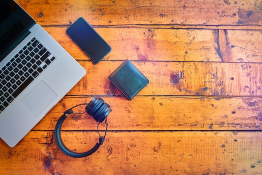 Top view of laptop, wireless headphones, mobile phone, and wallet on wooden table. Freelancer's work surface. Modern devices for business. Blank cell phone screen to text. Flat lay with copy space.