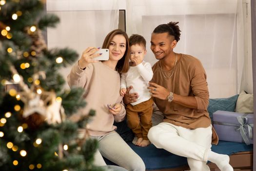 Happy young family in cozy sweatshirts: a girl with a small son, a stylish man, sitting at the window at home, taking a photo on a smartphone and congratulating friends on the Internet Merry Christmas