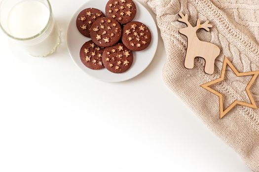 on a white table a saucer with round chocolate chip cookies, a glass of milk, a knitted beige plaid and wooden flat figurines of a deer and stars. View from above