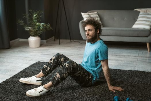 Handsome young man doing home work out sitting on the carpet relaxed at home looking at camera. Funny curly hair man with a mess beard doing exercise at home. Concept of healthy life.