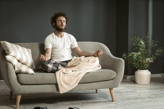 Young handsome man sitting on the couch or sofa at home enjoying meditation, relaxation, calmness, in his pajamas in living room during lockdown or being sick.
