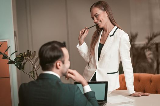 Young woman sitting on the table and sexually looking at young man. Two young and attractive colleagues man and woman flirting at office. Flirting in the workplace. Office romance.