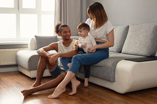 Happy, modern, multiracial family with a little boy in cozy home clothes sit on a gray sofa in romm, near a large window, smiling. Copy space