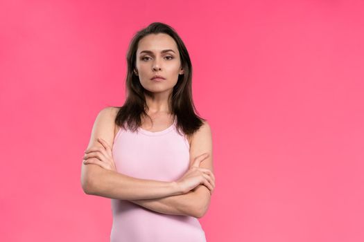 Suspicious young woman in a pink sleeveless t-shirt folded her arms and looks at the camera. Doubt, mistrust young beautiful woman isolated on pink background.