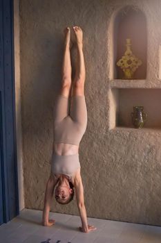 A beautiful and slender woman in a beige top and breeches, stands at a beige wall in her arms, upside down. Vertical