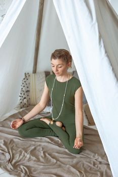 Beautiful and slender girl in green sportswear, meditates or practices yoga at home under a canopy of fabric, closing her eyes
