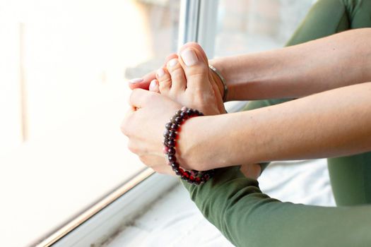 A slender woman in a green jumpsuit, in a yoga pose, clasped her feet in her arms, sits by a large window. Close-up