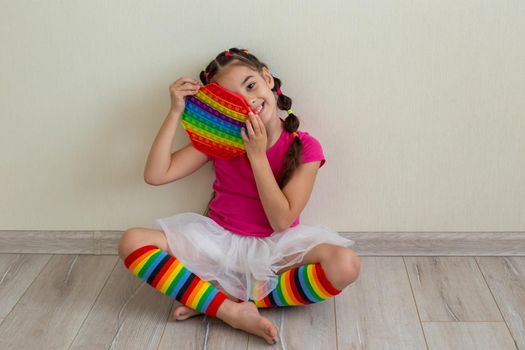 A cheerful little brunette in bright multicolored clothes and rainbow leggings sits on the floor against a light wall, holding a rainbow antistress toy in her hands. Copy space