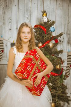 Delicate Girl of 10s in a White Fluffy Dress Holding a Big Christmas Gift near the Christmas Tree. Room is Full of Christmas Holiday Decorations. Close-up. High quality photo