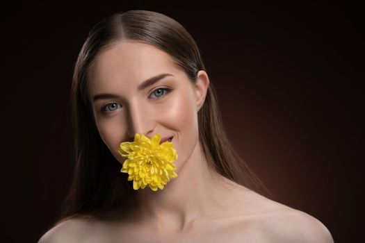 Smiling beauty model with bare shoulders holding a flower in her mouth with a beauty skin and long hair looking at camera isolated on black background. Woman nature cosmetic spa concept.