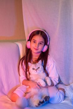 A beautiful little girl in pink headphones, with loose dark hair, sits with a white adorable cat in a neon pink-blue light