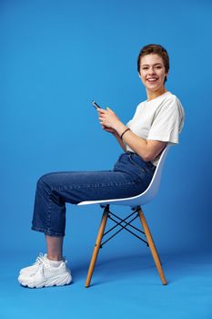 Cheerful attractive young woman holding smartphone, enjoying chatting or online shopping against blue background, close up