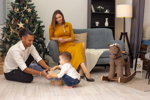 A beautiful slender girl in a mustard dress sits on a gray sofa, a young man and a toddler child cheerfully sort out boxes with gifts, on the floor, near a Christmas tree