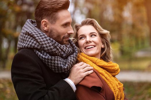 Close up. Happy in love young people, man hugging woman from behind when she looks at him, happy couple walking in a autumn park wearing stylish coats and picking up fallen leaves.