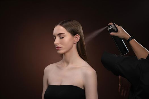 Hair stylist finishes work with model to apply hairspray to lovely girl hair. A beautiful model is getting ready for a show or a photo session with a makeup artist isolated on dark background.