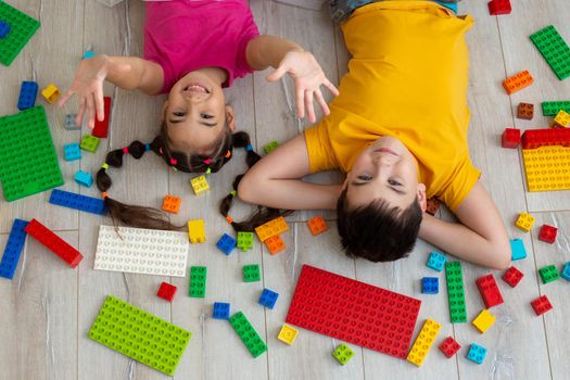 Portrait of happy boy and a little girl, brunettes, in bright multicolored clothes, lie on the floor, on their backs, in the room, with a multicolored plastic construction set