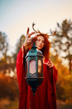 Woman in long red dress with deer horns in autumn forest. The demon of fairy tales in a red cloak with a lantern in his hands walks through the autumn forest and looks for magic herbs.