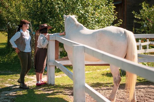 Happy farm living. Mother and daughter in vintage clothes stand near the paddock with a beautiful white horse. The girl is stroking a horse. Family at the ranch.