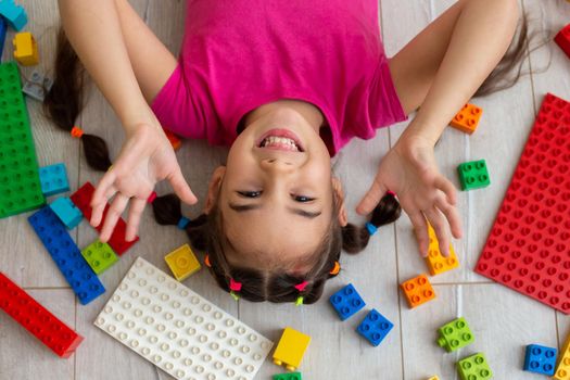 a cheerful little brunette little girl with two ponytails and multi-colored elastic bands for hair, lies on the floor, surrounded by scattered multi-colored parts of plastic construction sets.
