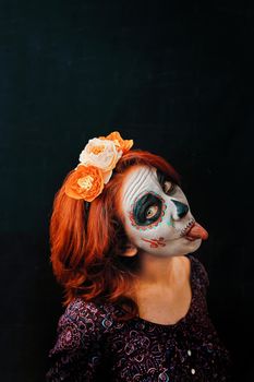 A young woman in day of the dead mask skull face art. Woman with skull makeup and red hair showing tongue on dark background, Halloween or horror theme