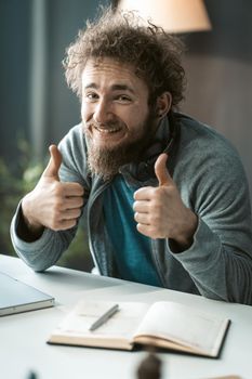 Thumbs up handsome satisfied young man in grey sweater and wild hair sitting working on laptop, looking at camera. Young bearded man, study at home, working on laptop smiling, and showing thumb up.