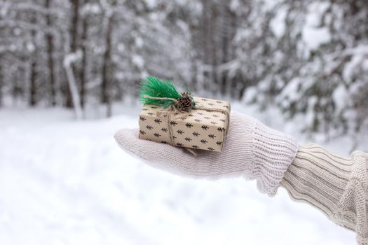 The girls hand in white mitten are holding a little gift box against the background of a winter forest. Close-up