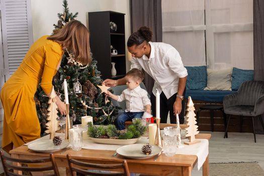 A young family is decorating a Christmas tree in the living room. near a tree, a man with a small son hang a wooden figure of a star. Mom in a mustard dress stands in front of the fir tree, helps.