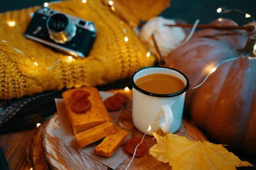 Autumn holiday composition for postcards, invitations and posters. Tea, carrot cake, fall leaves, knitwear, vintage camera and lights from garlands. Log of a tree as a tray.