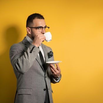 Handsome young bearded business man in eye glasses in formal wear holding a cup of coffee standing sideways isolated on yellow background.