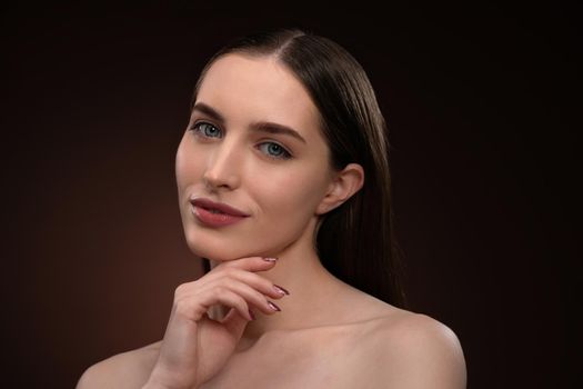 Beautiful spa model girl with perfect fresh clean skin and bare shoulders. Charming brunette model girl posing on camera touching her chin with a finger isolated on black background.