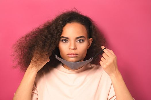 No emotion or indifference holding reusable mask African American girl in peachy t-shirt, to prevent others from corona COVID-19 and SARS cov 2 infection isolated on pink background.