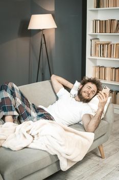 Sick young man in pajamas sleeping on the couch looking at smartphone or having video call . Handsome bearded young man sleeping on the sofa after a long hours working late nigh.