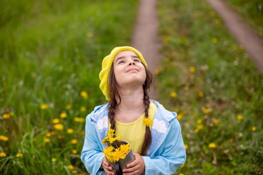 A little girl with two pigtails, in a yellow beret, a blue raincoat, stands in nature on a green lawn with dandelions, looks up, holds a bouquet of yellow dandelions