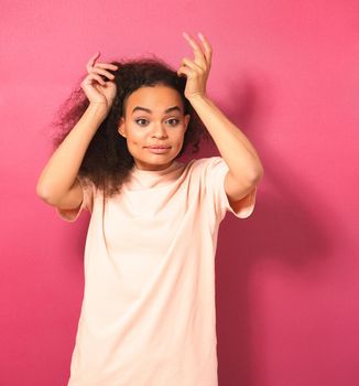 Beautiful young African American woman with Afro hair dancing looking positively at camera wearing peachy t-shirt isolated on pink background. Beauty concept.