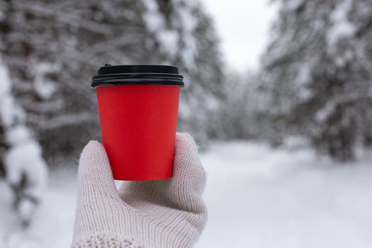 Red paper cup of coffee in hand in a white knitted mittens, against the backdrop of a beautiful winter forest. Copy space