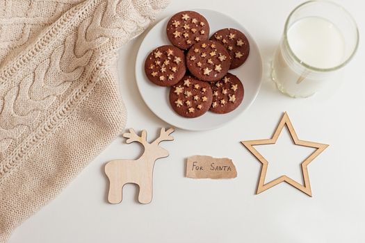 on a white table a saucer with round chocolate chip cookies, a glass of milk, a knitted beige plaid and wooden flat figurines of a deer and stars, and a piece of paper with the words -For Santa. View from above.