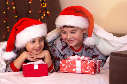 Two cute children, a girl and a boy, in pajamas and red hats, cuddle on the white bed with gifts in their hands.