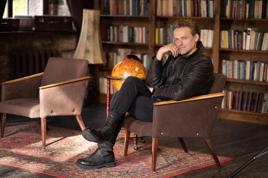 pensive man in a leather jacket and ankle boots sits cross-legged in an old armchair in a vintage room. Caucasian man sits in an armchair in the middle of an antique living room against the backdrop of shelves with books. Rustic rug on the floor. Close-up . High quality photo
