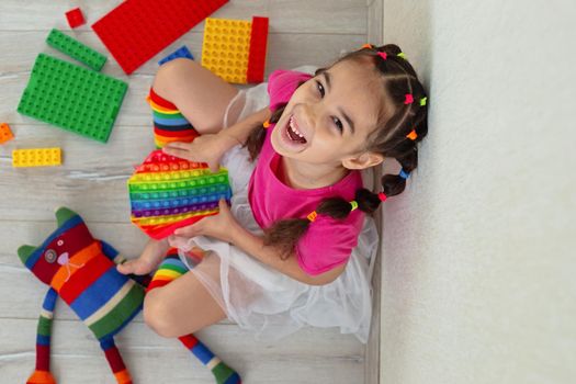 cheerful little brunette girl in bright colorful clothes, sits at home, on the floor, surrounded by colorful toys and rainbow pop it. Looks at camera. View from above