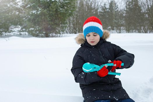 A teenage boy in a black jacket and a striped knitted hat, sits in the snow, makes snowballs using a light blue plastic sculpting tool. Close up