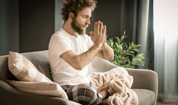 Serene Man Meditates in lotus Position on His Couch At Home. A Man With Folded Hands Prays in the Morning. Close-up. High quality photo