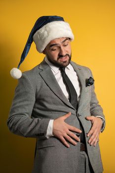 Overeat young handsome bearded man touching his belly wearing blue Santa Claus hat and suit, looking at camera isolated on yellow background. Positive emotions, feelings seasonal holidays.