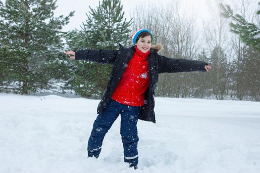 A cheerful teenager in a dark jacket and a red sweater stands in a winter park on the snow against the backdrop of young pines