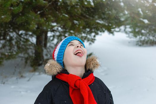 Funny boy in winter clothes and a red scarf walks during a snowfall, catches snowflakes with his tongue